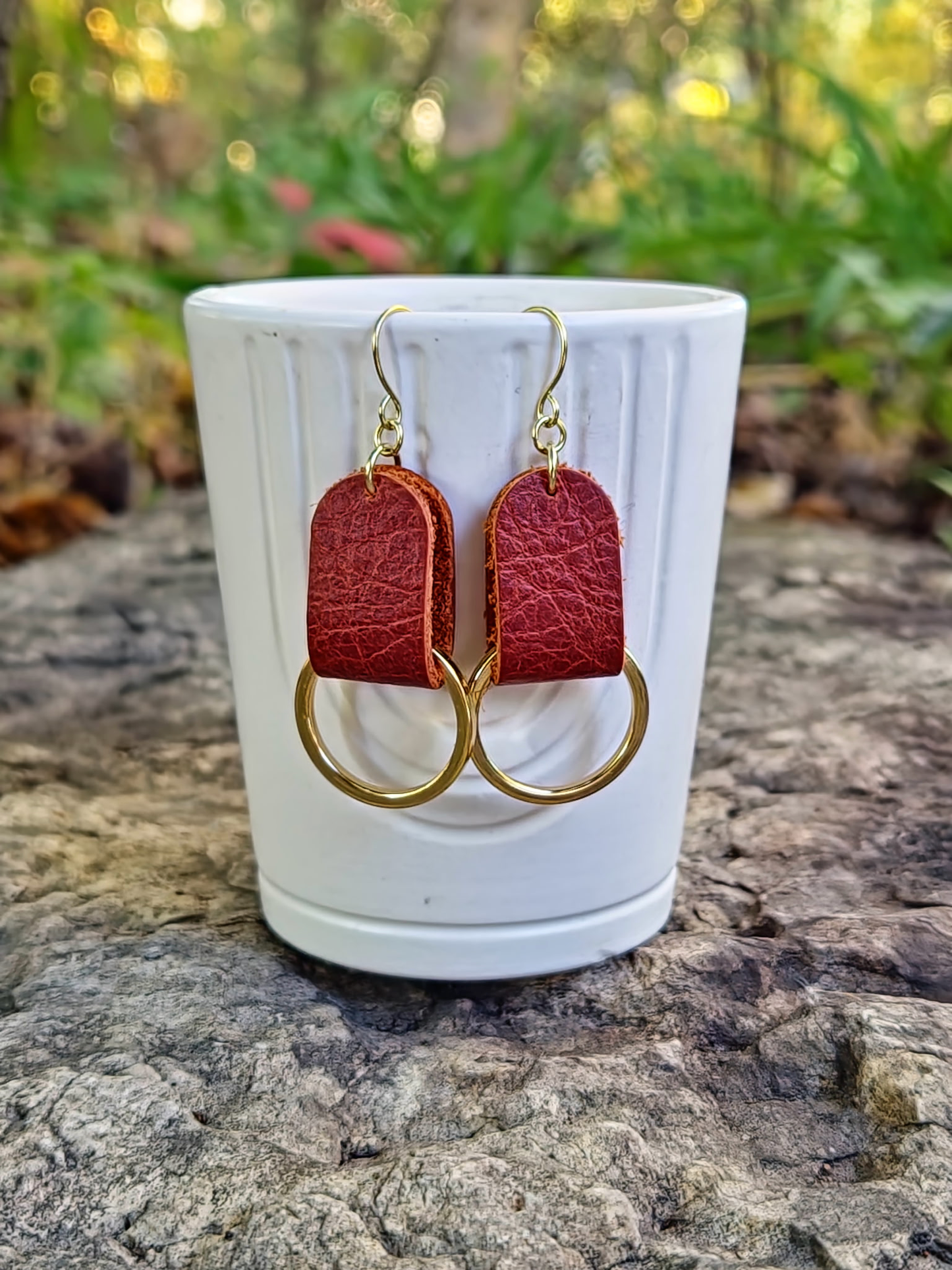 Rusty red leather earrings with gold loops on a white cup against a natural background